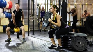 Woman in gym performs box squat holding weight plate, man trains using a kettlebell next to her