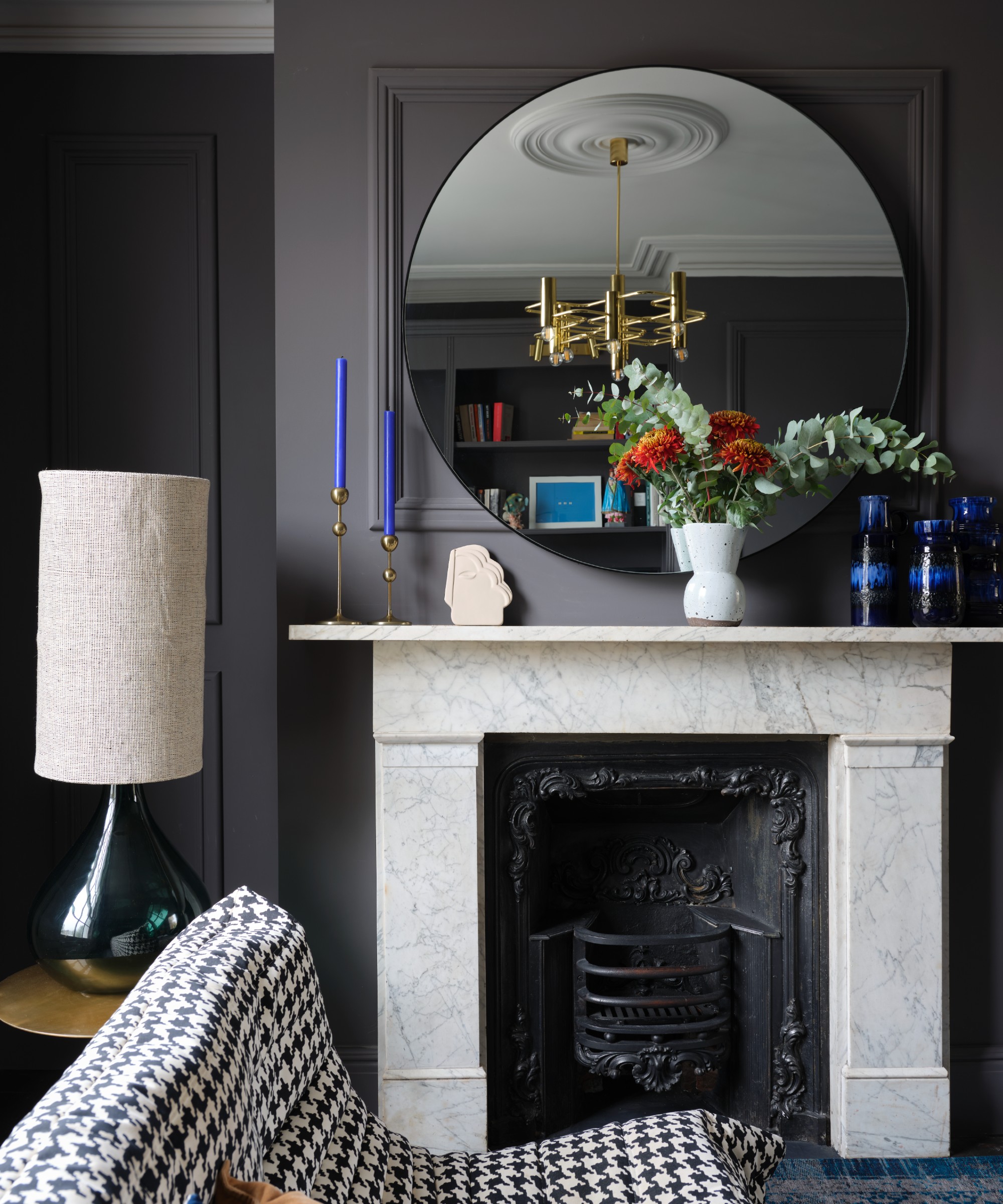 A living room painted in dark grey with a marble fireplace, a round mirror and a houndstooth-print settee