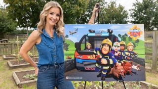 Helen Skelton poses on a farm next to an easel with the key art from Fireman Sam displayed on it