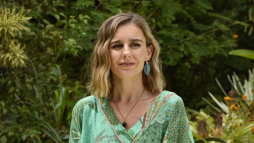 Susie Montagu (Georgia Maguire) stands with lots of lush green vegetation in the background behind her. She is wearing a V-neck green desk with floral prints on it and a chain around her neck. She is smiling softly.