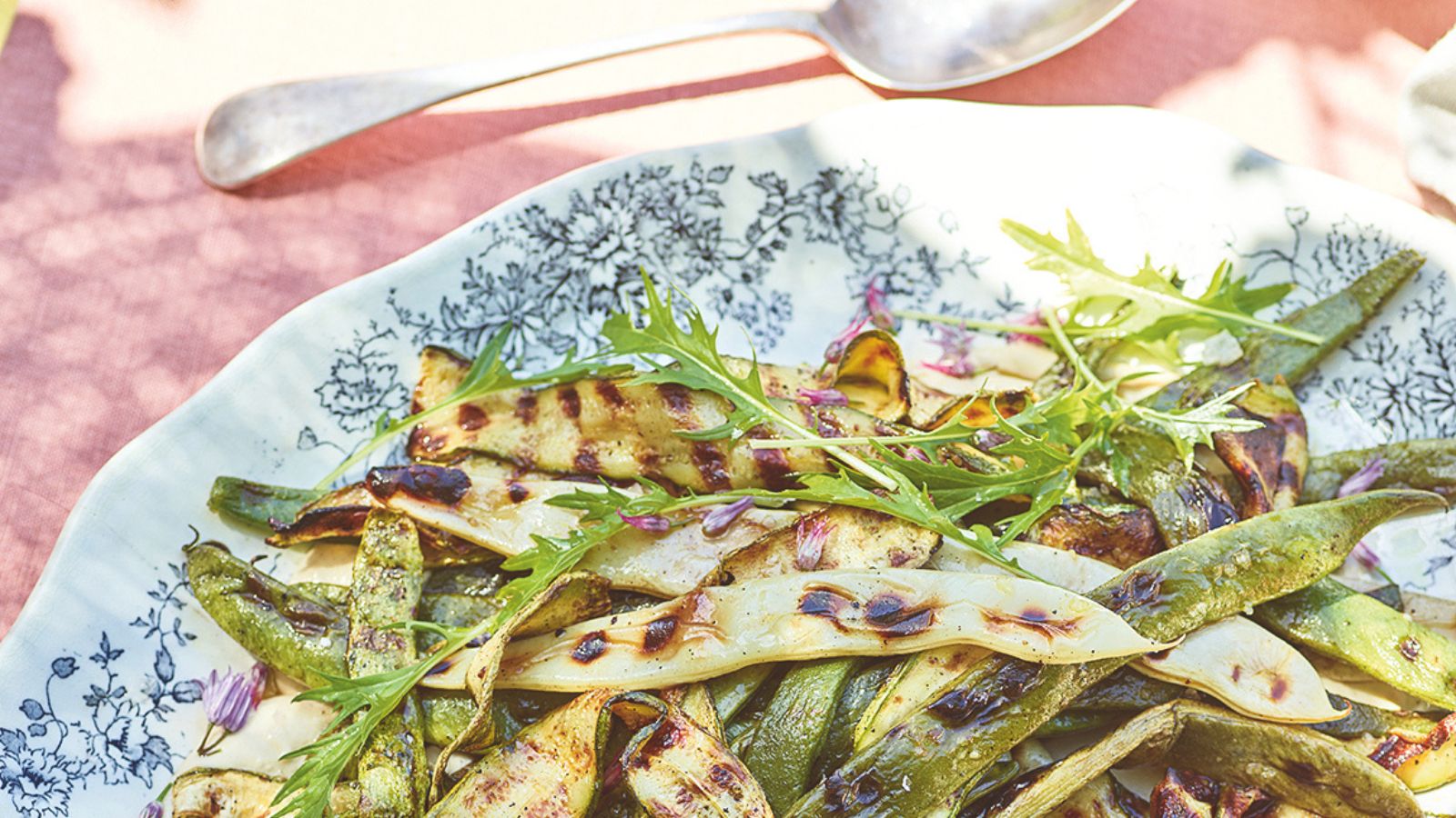 Platter of griddled courgette and beans in a tahini sauce on a floral platter over a pink tablecloth in the sun