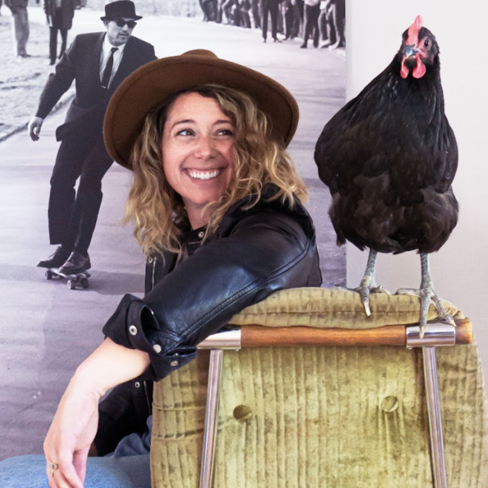 A picture of Lisa Carolla, a woman wearing a brown hat wearing a leather jacket, smiling to the right at a black chicken standing on a woven box, with a black and white print of a man behind her