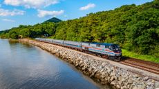 A train on the Empire Service route chugs along past a lake and green trees on a sunny day