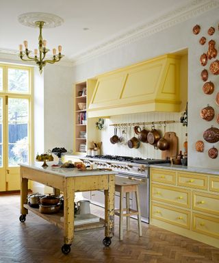 Large, traditional white kitchen with bright yellow kitchen units and painted window frame, wooden kitchen island with wheels, dark parquet flooring, walls decorated with traditional metal pans, metal chandelier
