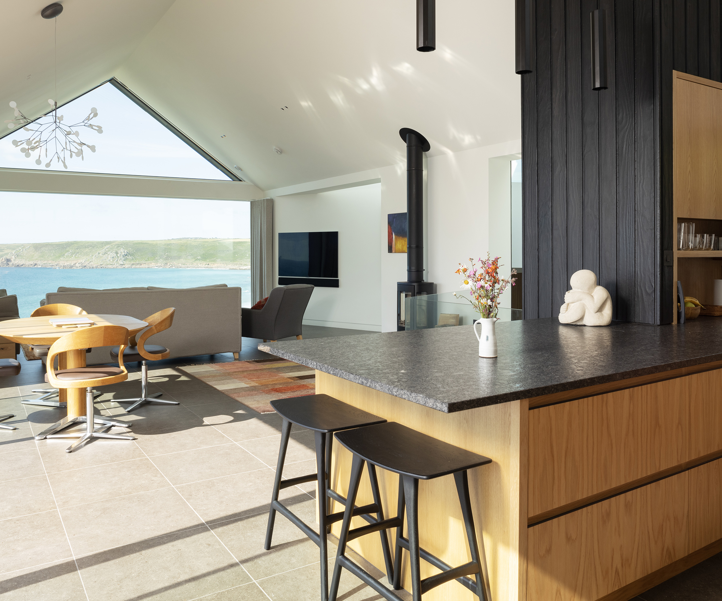 In the foreground there is a wooden kitchen with black work surface on a peninsula with bar stools. The background looks out to an open plan dining and lounge area with huge picture window
