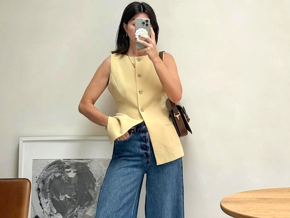 British influencer Francesca Saffari poses for a mirror selfie in her home wearing a butter-yellow button vest top, brown leather bag, and wide-leg jeans