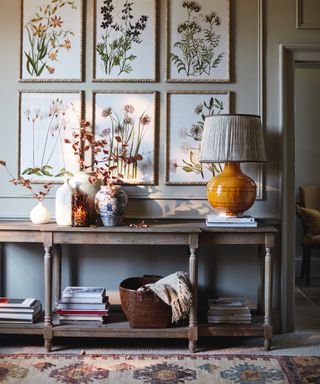 how to design a home that feels like you, entry way with botanical prints on stone coloured wall, mustard lamp base with neutral shade, vases, large console with shelf, books on shelf, basket, antique style rug