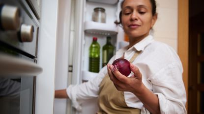 Woman wearing a white shirt and a brown apron stands in front of open fridge holding a red onion