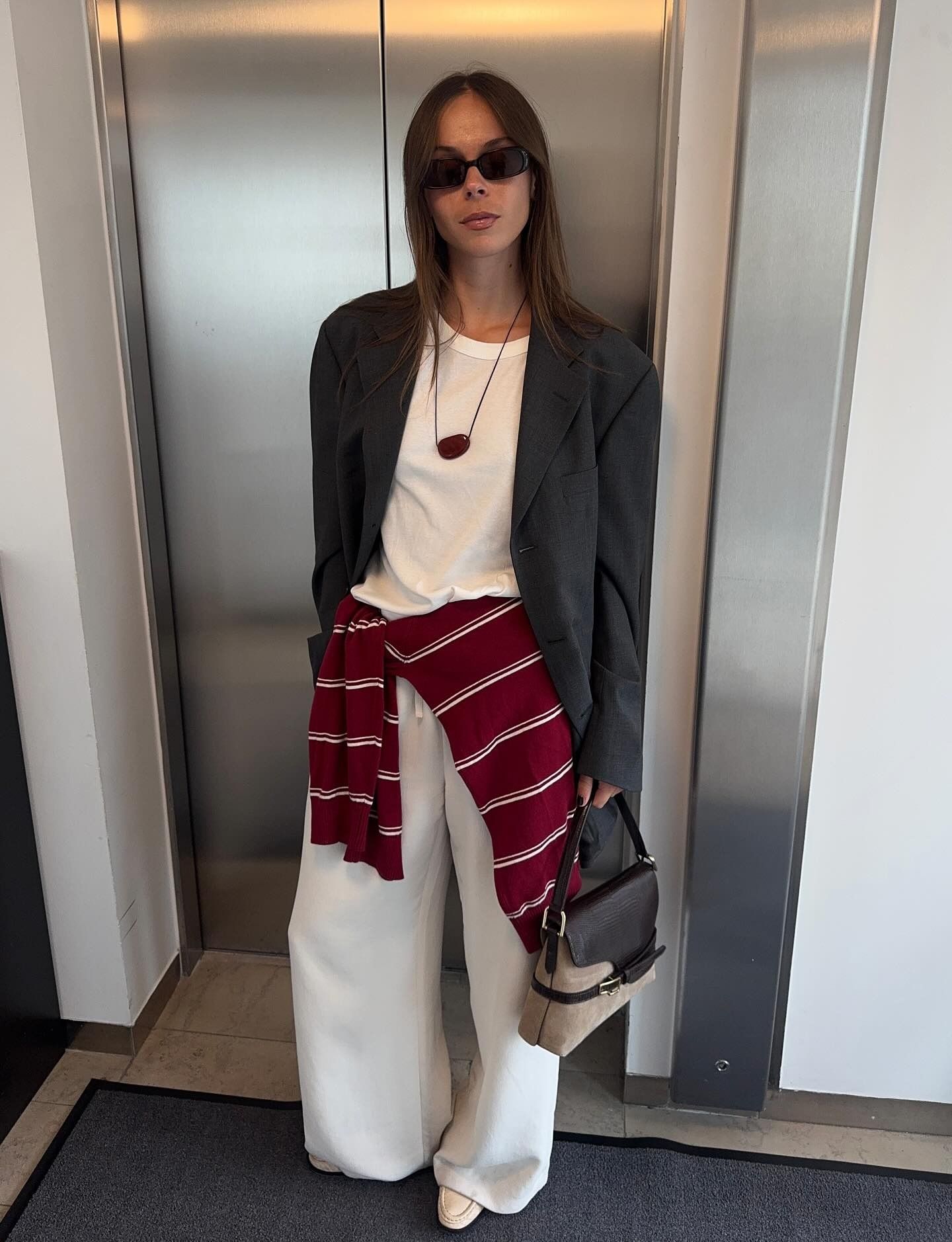 Swedish fashion creative Johanna Lager poses in a chic fall outfit with a gray blazer, cord necklace with a red pendant, a white T-shirt, a striped red sweater tied around her waist, and baggy white jeans