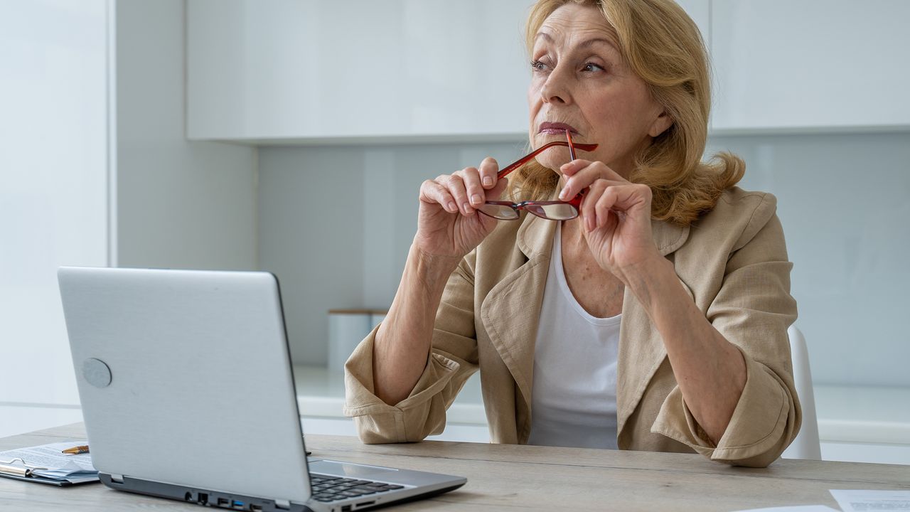 An older woman looks thoughtful while she sits in front of her laptop in the kitchen and looks into the distance.