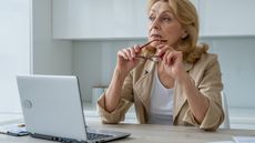 An older woman looks thoughtful while she sits in front of her laptop in the kitchen and looks into the distance.