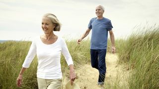 A smiling senior woman wearing a white top and her husband dressed in a blue shirt walk across sand dunes