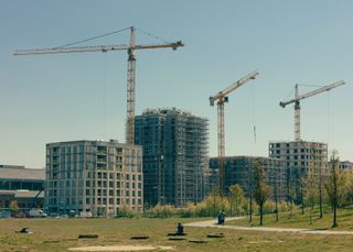 A row of buildings being constructed with cranes above them and a park in front of them.