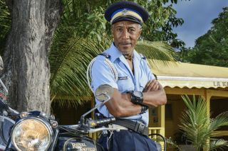 Danny John-Jules in character as Officer Dwayne Myers, in uniform and leaning against a motorcycle with his arms folded while standing under a palm tree