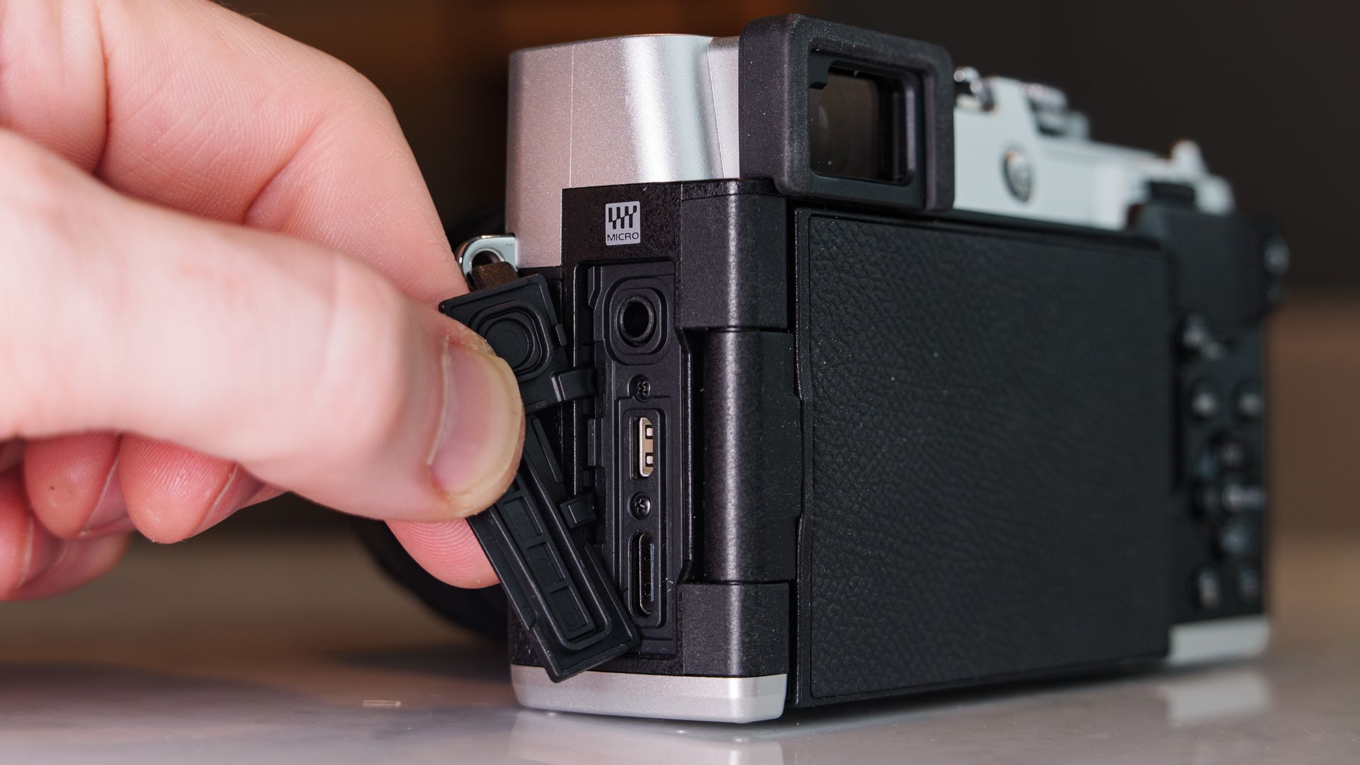 Close up of the OM System PEN mirrorless camera's connection port doors, they are being opened to reveal the ports, in a modern room with lantern in the background