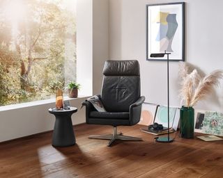 Rich tone, textured wooden flooring, with dark leather swivel chair, staggered artwork, small round side table, and pampas grass in floor vase.