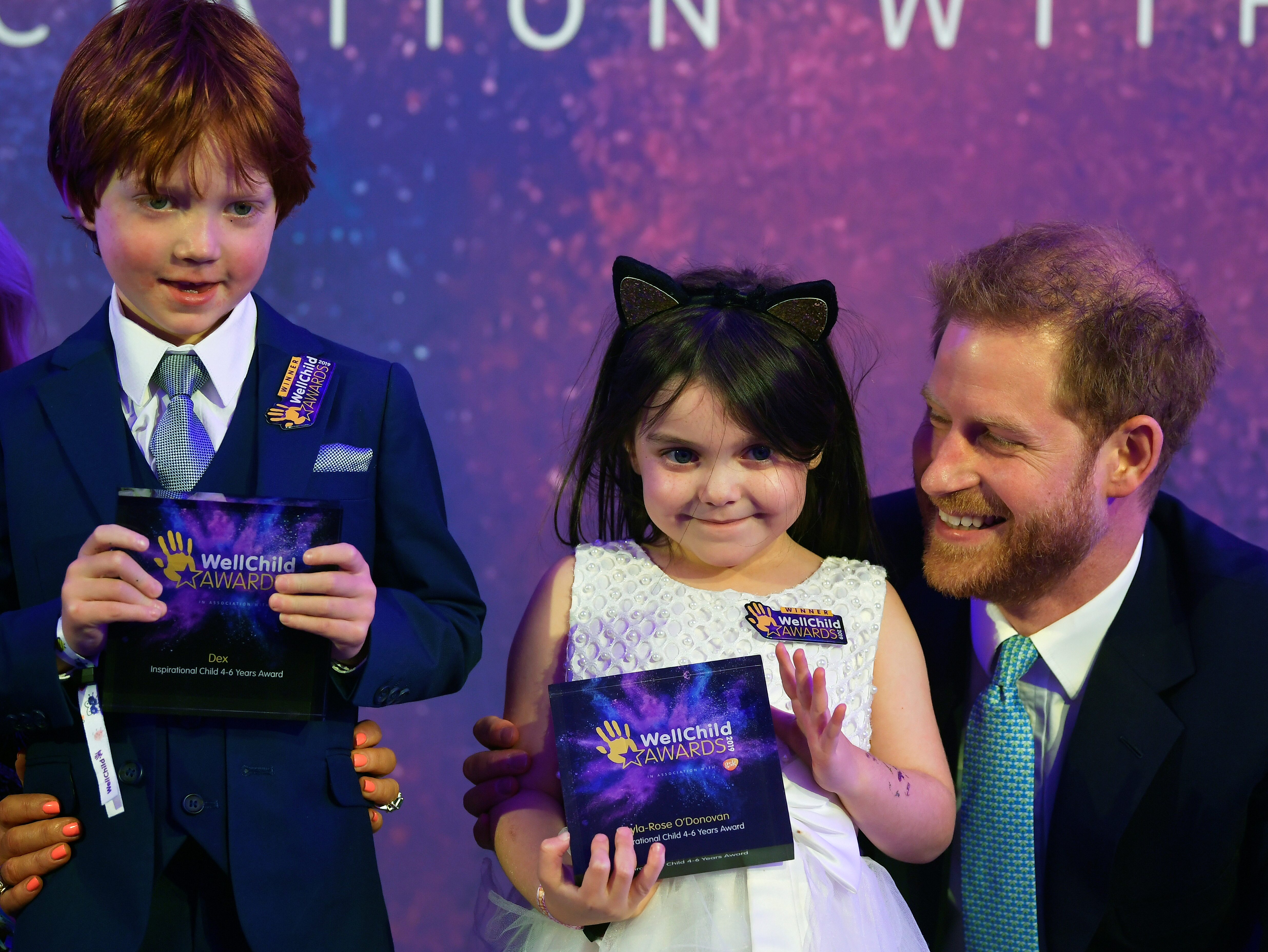 Prince Harry smiling and kneeling next to two young children holding awards