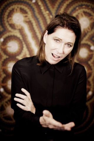 A portrait of Kim Deal gesturing at the camera in front of a patterned background