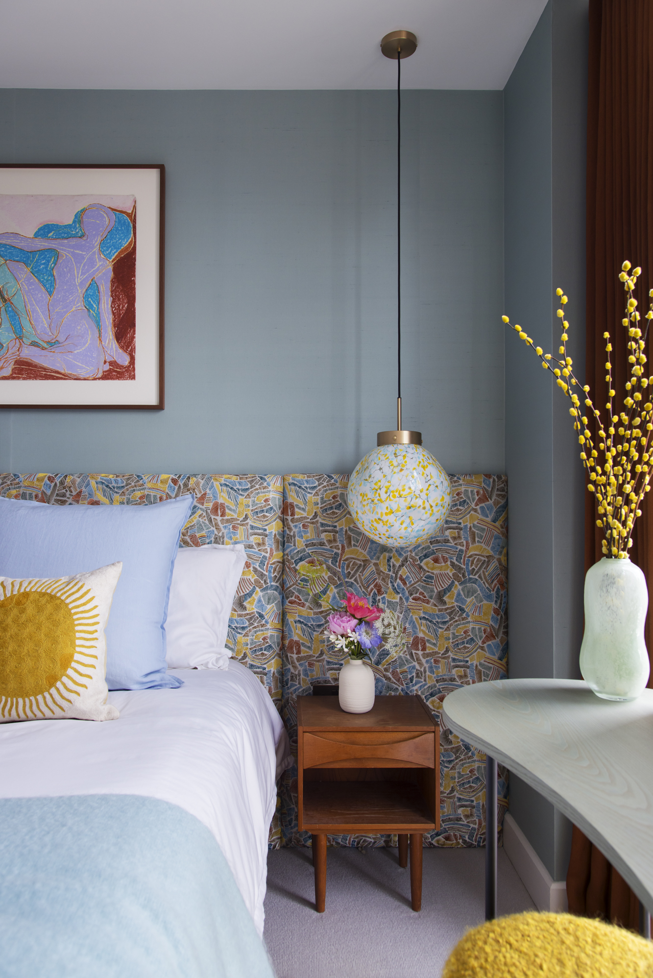 modern bedroom with blue wall, colorful headboard, blue and white sheets with yellow pillow, floating orb pendant light, timber side table, and a console table