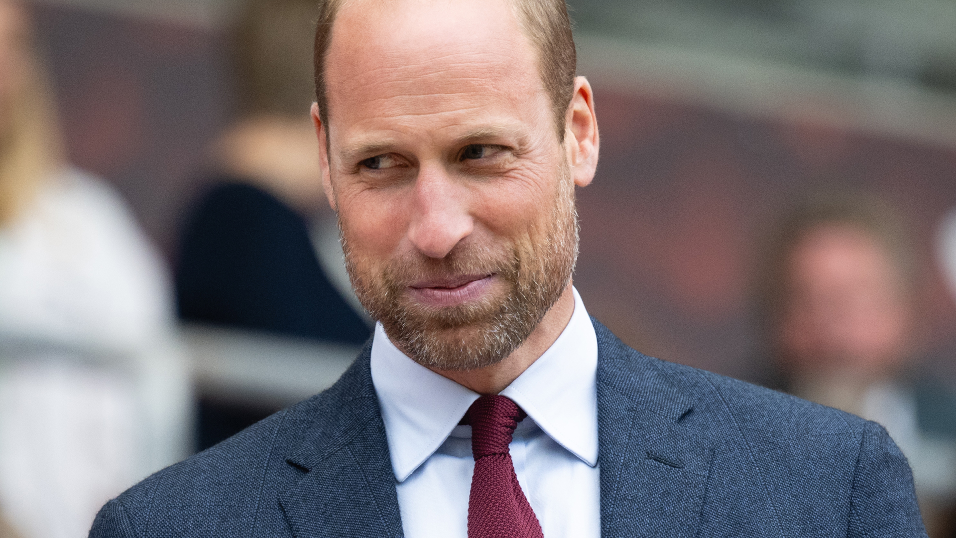 Prince William smiling while wearing a dark gray suit, a dark red tie, and a white shirt