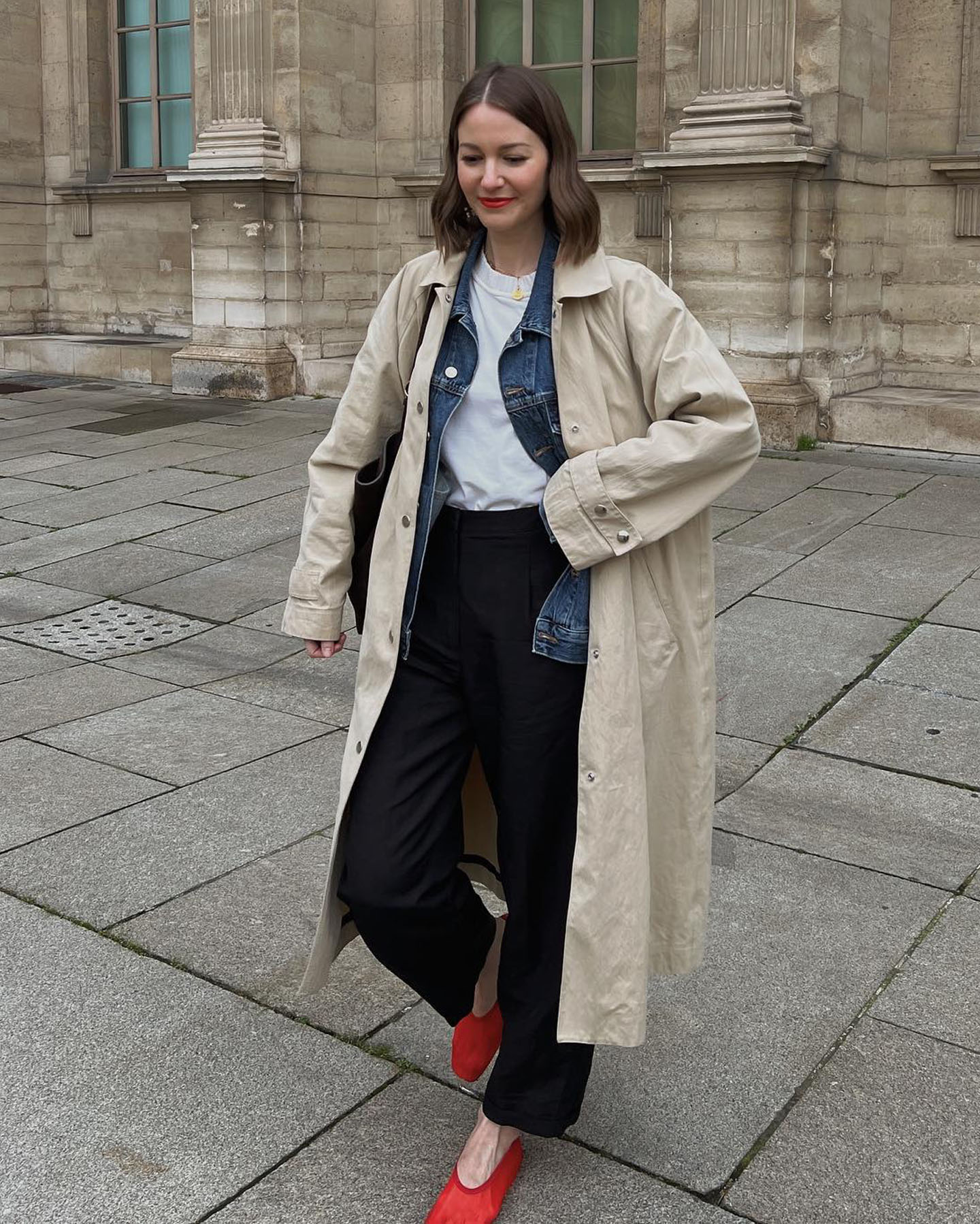 fashion influencer Marissa Cox poses on the sidewalk in Paris wearing a trench coat, denim jacket, white tee, black pants, and sheer red mesh flats