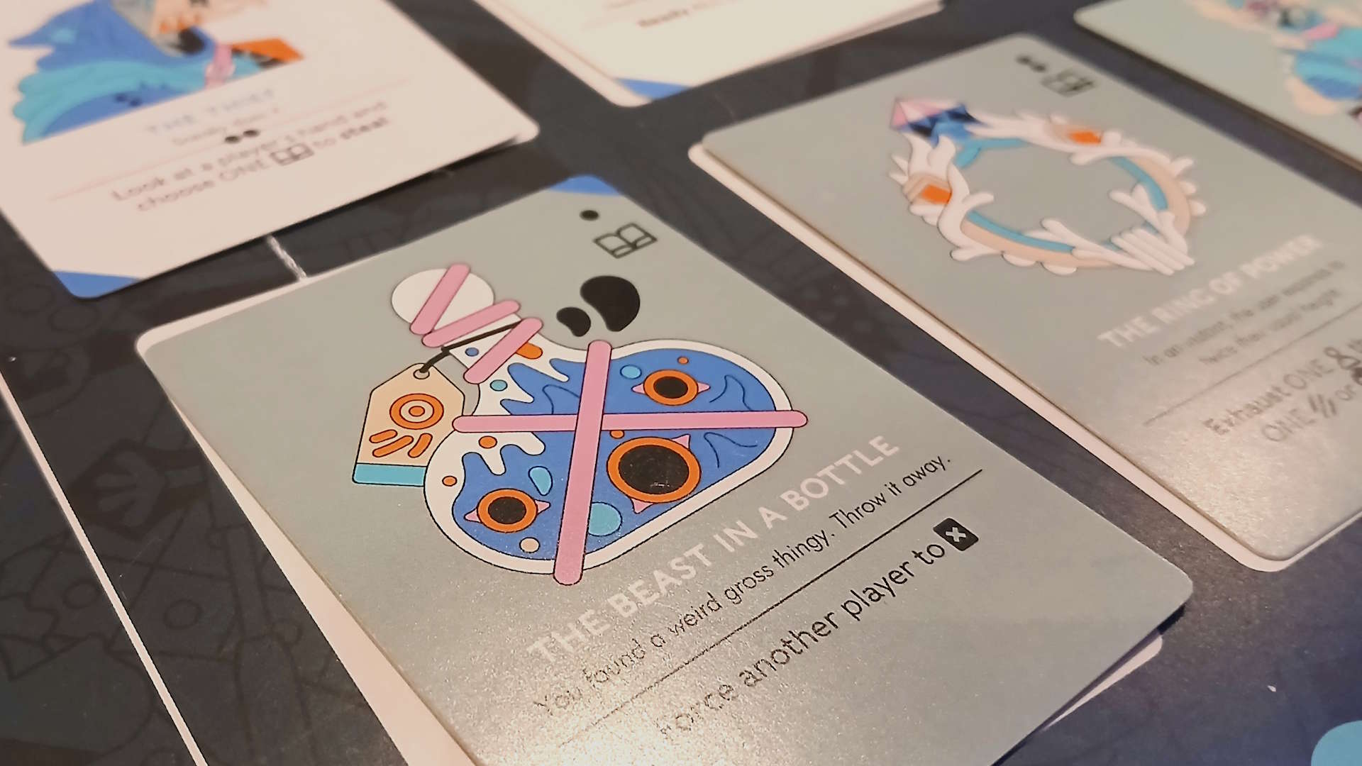A series of cards laid out on a dark surface, with one card reading &#039;The Beast in a Bottle&#039; with a cartoon bottle on it taking up much of the foreground
