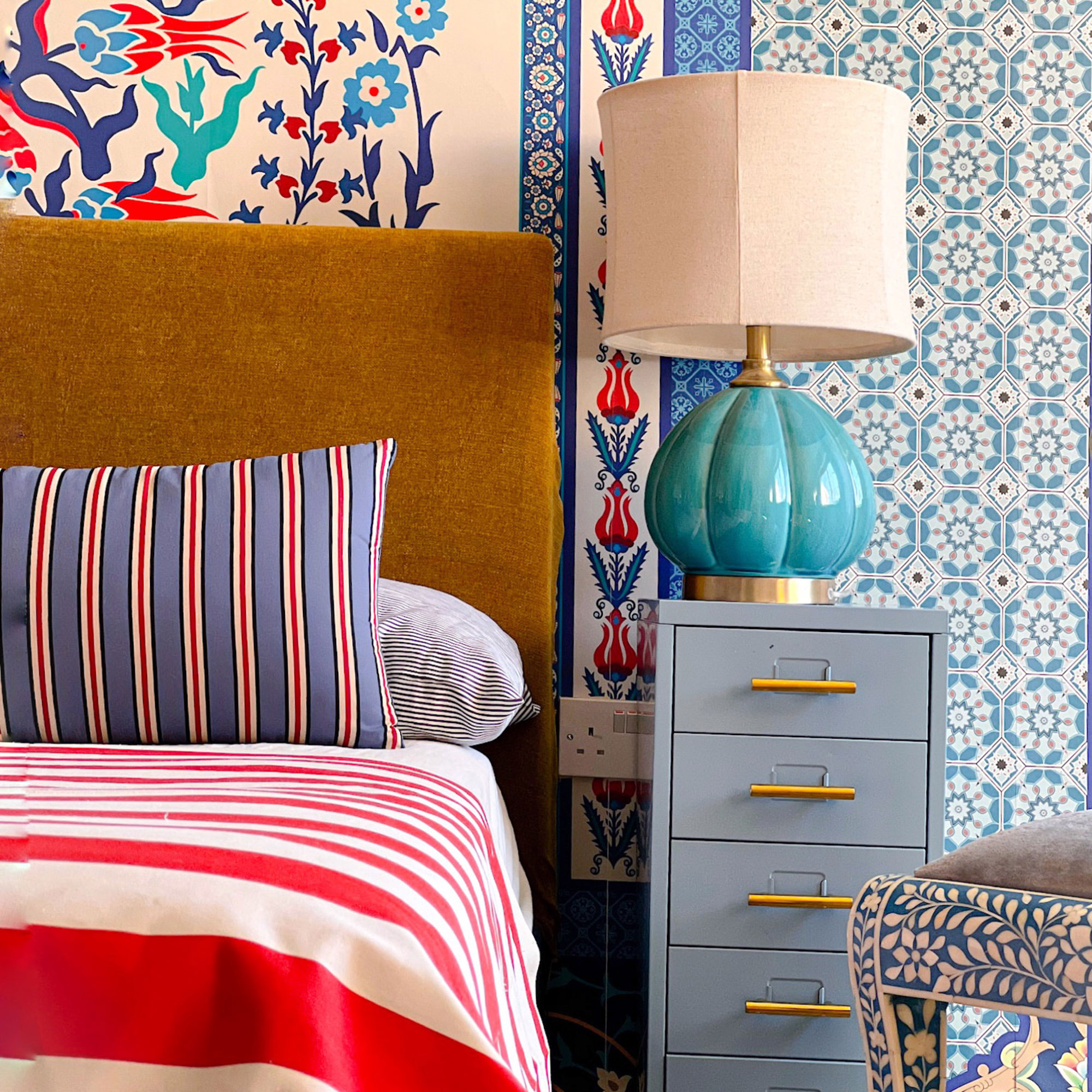 Bedroom with ochre upholstered headboard, patterned walls, pattern mix and matched pillow and bed covering beside pale blue-green storage unit with turquoise bedside lamp