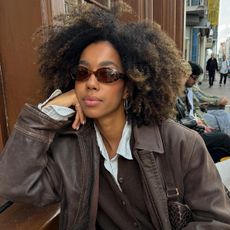 Influencer wearing button down shirt, brown sweater, and brown leather jacket while wearing brown oval sunglasses and sitting on outdoor cafe bench.