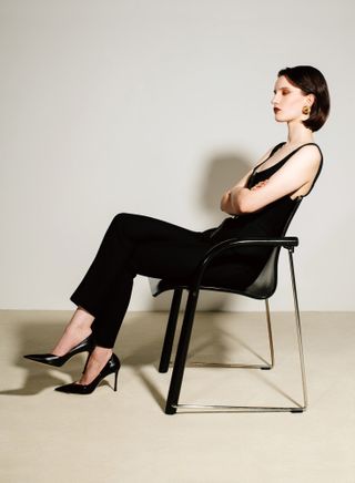 Model sits in chair, wearing black Hermès jumpsuit