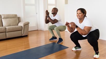 woman and man performing squats in a living room setting on a wooden floor with a grey exercise mat in front of them and a sofa to the side.