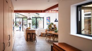 A wide shot of the interior of a kitchen extension. There is an interior courtyard to the right of the shot which can be viewed out of a window. The beams are exposed and there is a female figure standing at the glazed doors looking out to the back garden