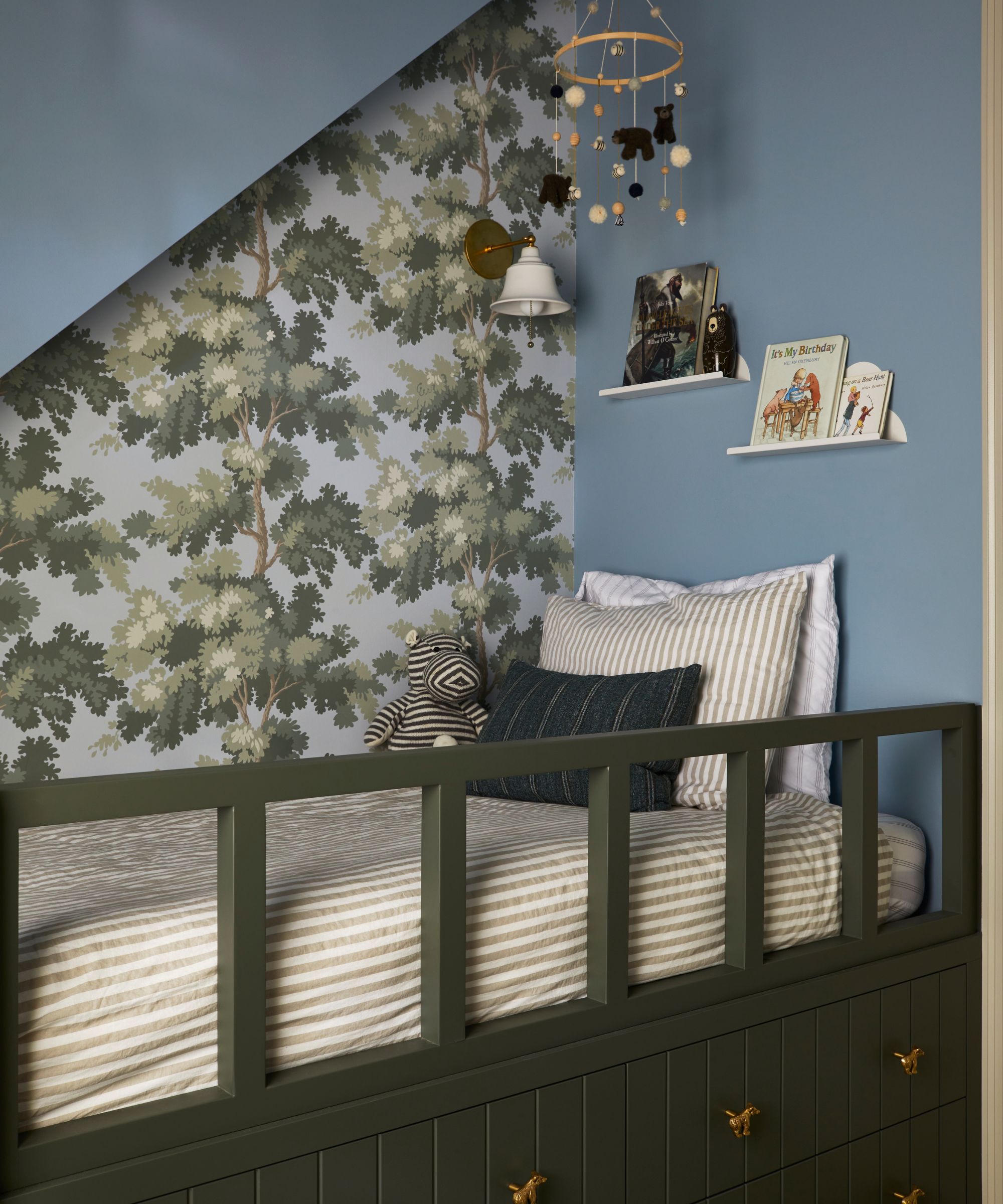 a little boys bedroom with a blue painted sloped roof, a green custom bed with railing and storage beneath with a tree patterned wallpaper