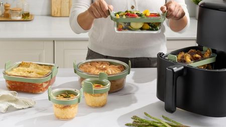 A set of glass air fryer dishes with green silicone rims, on a white counter beside a large black basket air fryer. 