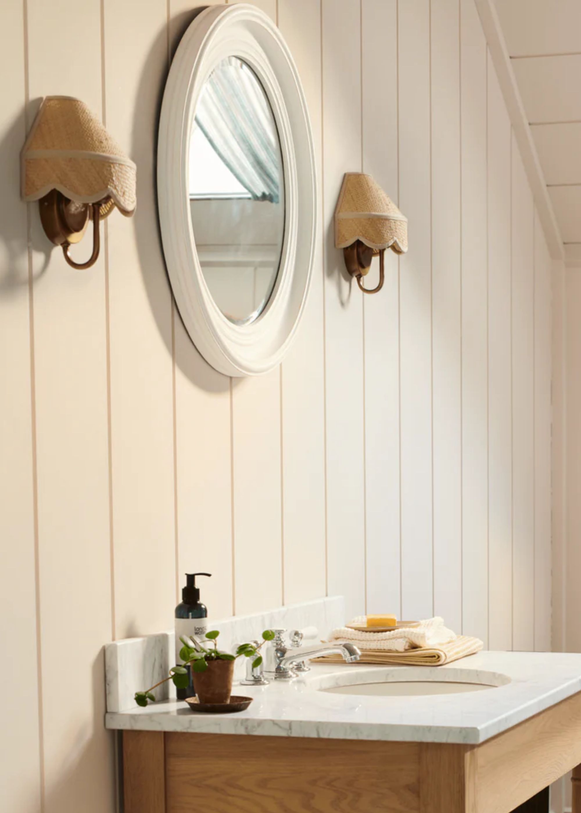 A small bathroom with two wall lights hanging next to an oval shaped mirror. There is also a basin with silver hardware and bathroom essentials on top of the counter.