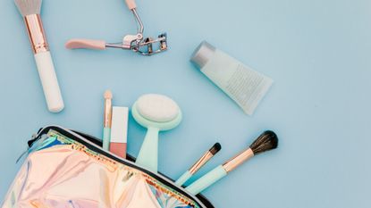 A makeup bag with makeup and brushes falling out on a blue background