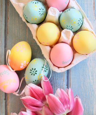 Six pastel patterned colored eggs - mint, yellow, and pink - in an egg crate, with three eggs on the distressed light blue wooden table and pink tulips below them