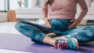 Pregnant woman shown in exercise clothes and sneakers, holding her stomach and sitting on an exercise mat in her home 