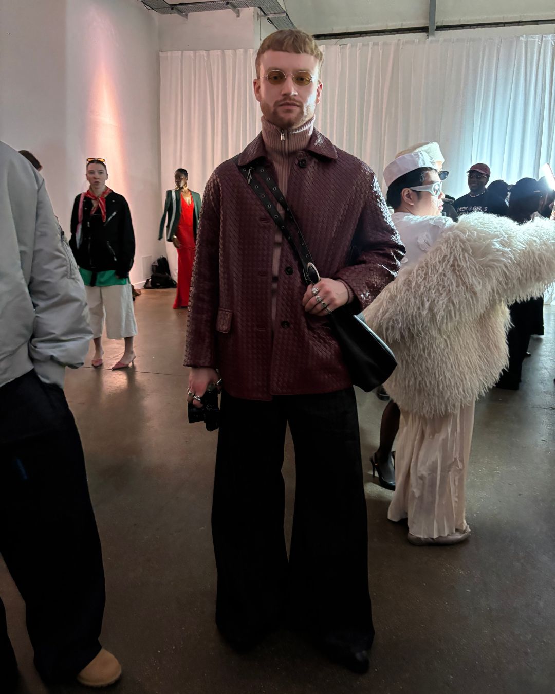 An image of Jonathan Vine, a fashion influencer (@vine.fits) at London Fashion Week, wearing wide leg / flared black trousers with a woven-textured burgandy-brown jacket, over a light red high-neck zip up jumper