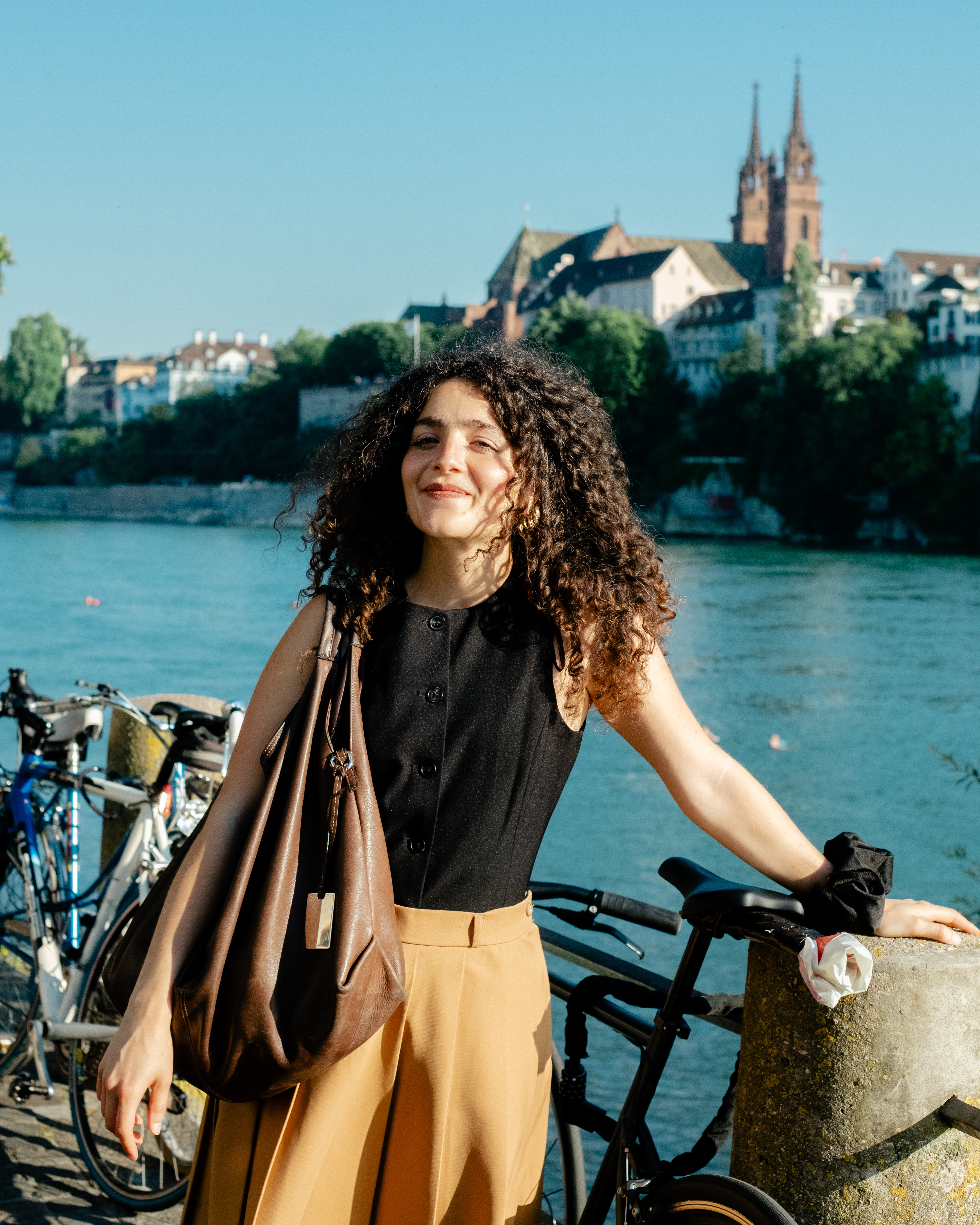 A ypung woman dressed in a black bottoned-up top and a beige, pleated skirt smiles at the camera while standing in front of a river canal bathing in sunshine, while in another image, two middle-aged people sit on a bench on the same, leafy canal.