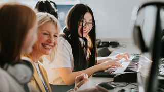 A kind female teacher in a yellow blazer takes some time to talk to two female students individually. She crouches down between them as they sit in a music / media studies classroom. 