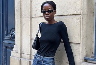 Influencer wearing black long sleeve top, black sunglasses, and straight-leg jeans while leaning up against Paris wall.