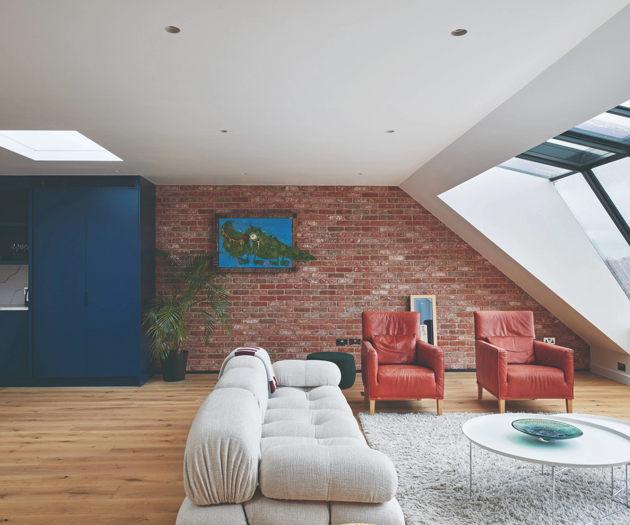 Loft conversion with red chairs, cream sofa, red brick wall and windows to the right and blue kitchen to the left