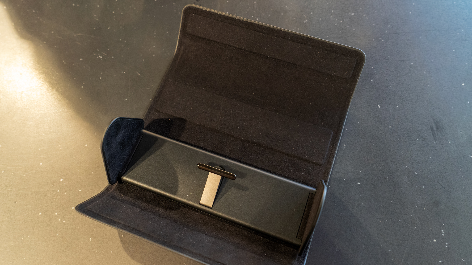 The charging case for the Meta Ray-Ban Display glasses opened out on a table is black and at the base a center piece which would sit between the frames is visible. There is a battery block beneath.