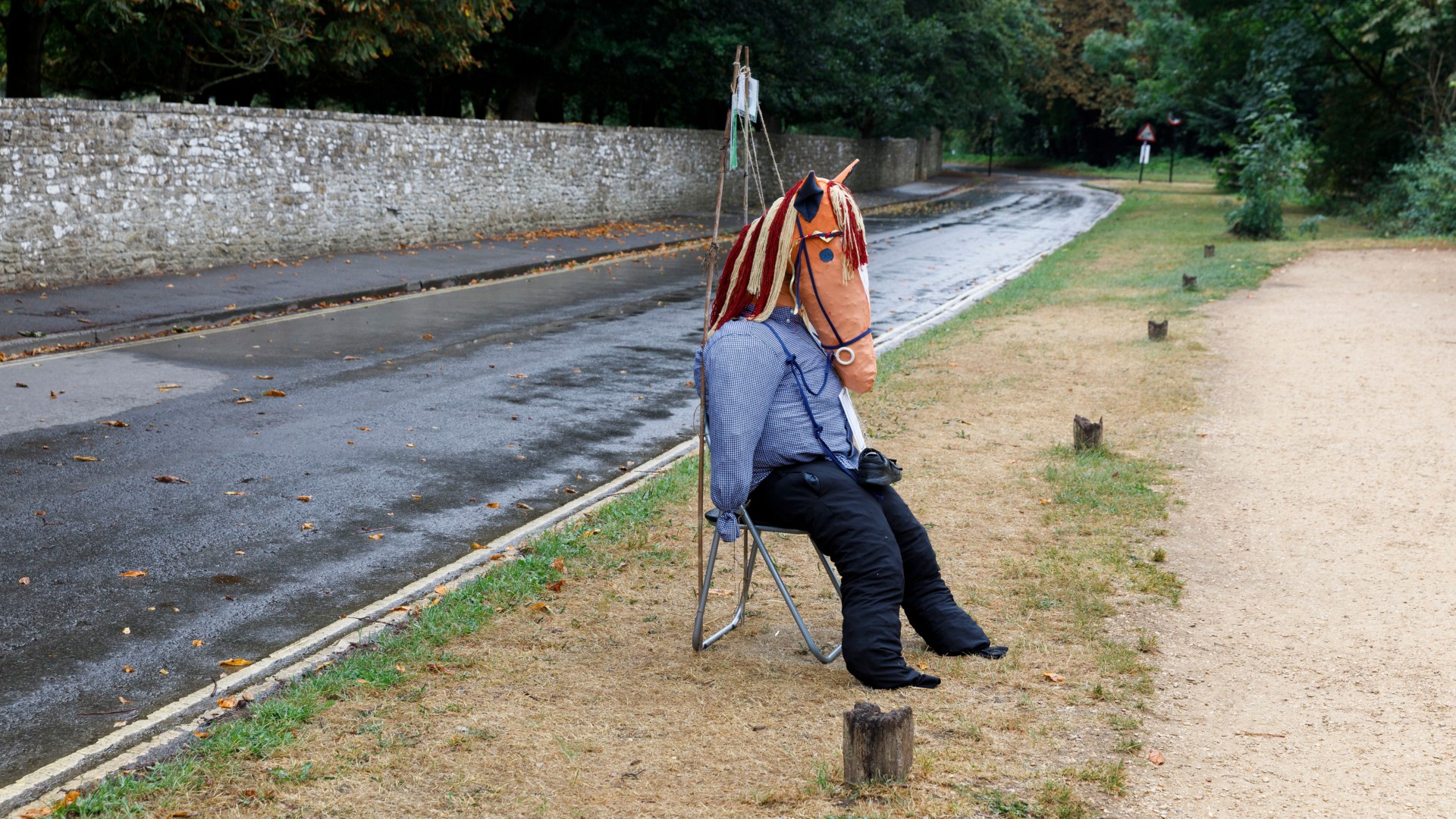 A scarecrow dressed in a blue checked shirt and black trousers sits on a folding metal chair at the edge of a quiet, wet road lined with trees and a stone wall. Its head is a handmade fabric horse's head with a rope mane, and a stick supports it from behind.