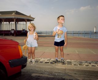  A photograph of two children eating ice cream, standing beside a red car
