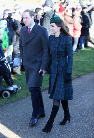 Kate Middleton wearing a green-and-blue plaid coat and black boots holding hands with Prince William walking p ast a crowd of people outdoors