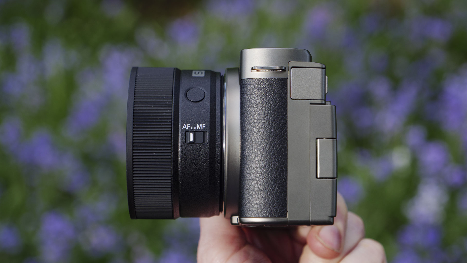 Side profile of the Panasonic Lumix S 40mm F2 lens, attached to a 'titanium' version of the Lumix S9, with natural bluebell backdrop