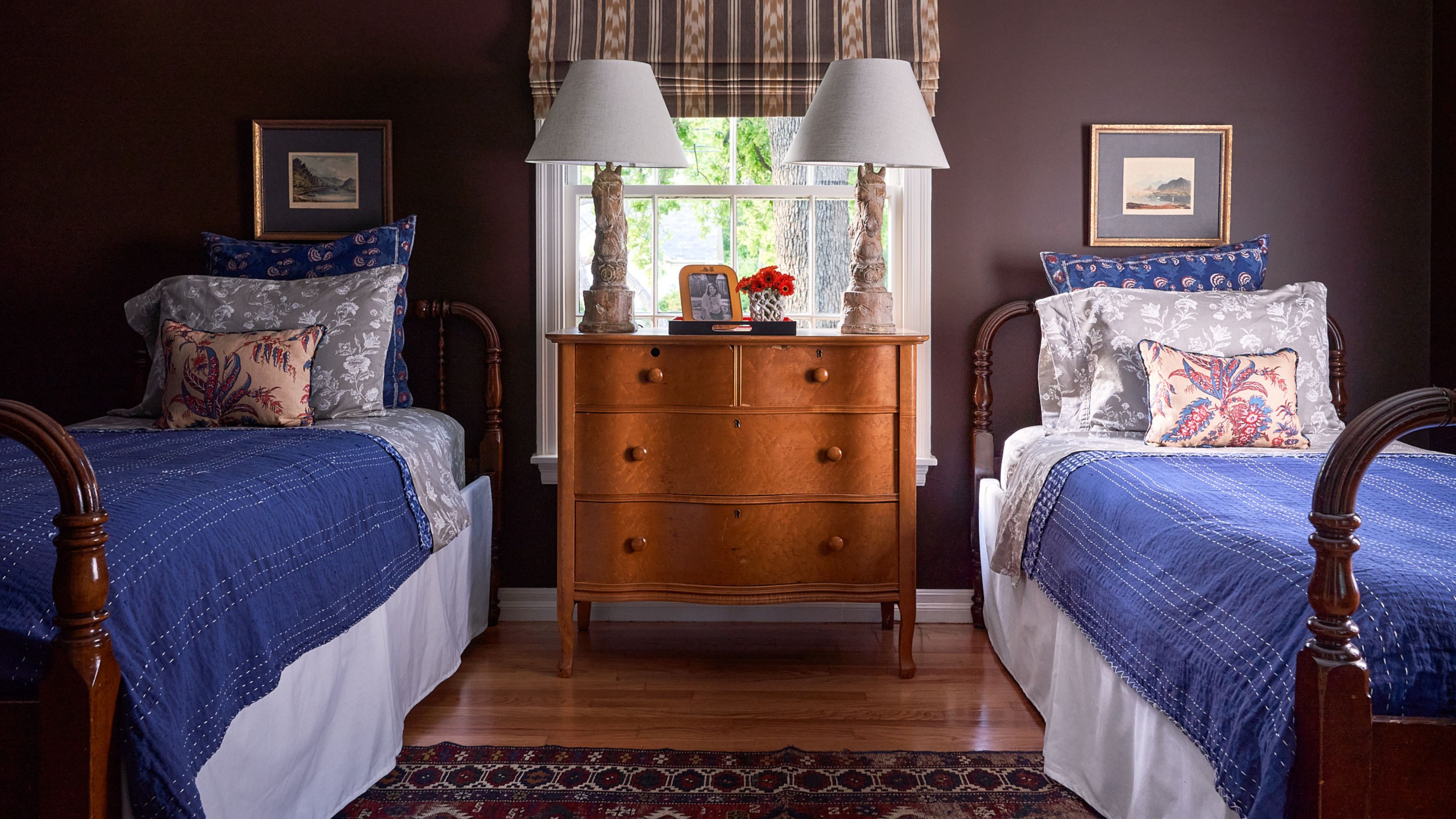 a dark purple burgundy bedroom with two antique twin beds, layered blue linens and antique dresser and lamps in front of the window