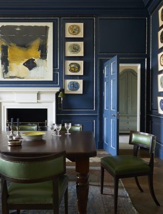 A dining room painted blue with antique artwork, a wooden table, and green dining chairs