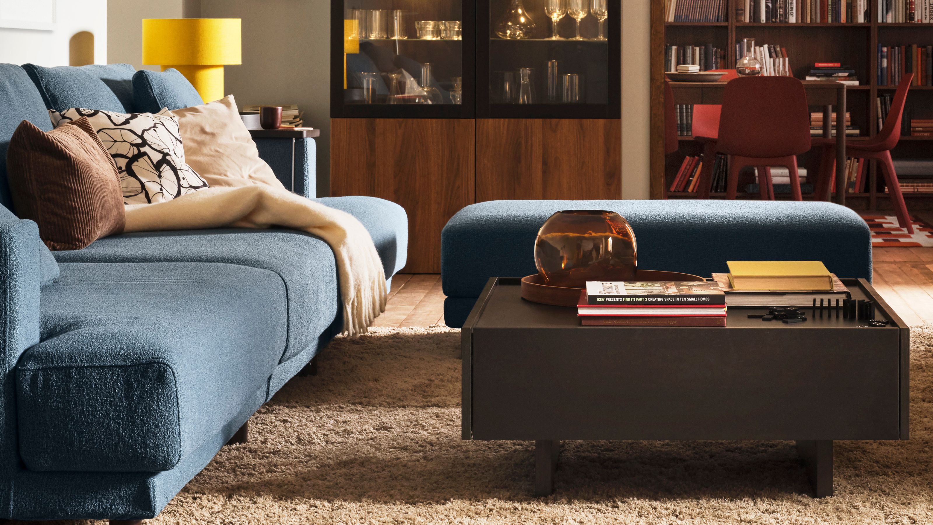 A modern living room with a square brown coffee table, a brown sofa and pouf, a side table with a yellow table lamp, and glass and open shelving behind with a dining area tucked into a nook